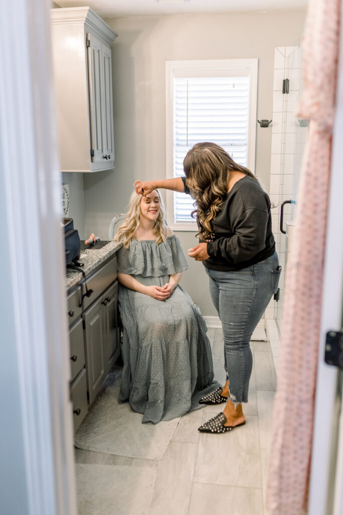 Mom in green dress in her own home having hair and makeup done by hair and makeup artist in her own bathroom before in home newborn session. 