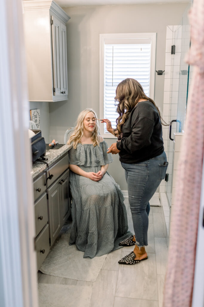 Mom in green dress in her own home having hair and makeup done by hair and makeup artist in her own bathroom before in home newborn session. 