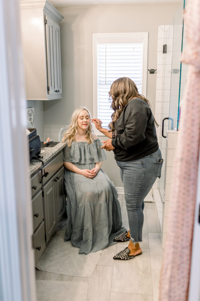 Mom in green dress in her own home having hair and makeup done by hair and makeup artist in her own bathroom before in home newborn session. 