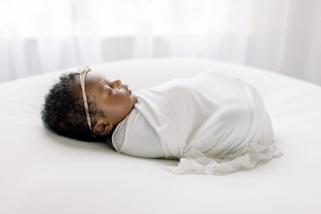 Newborn baby girl in studio swaddled in white fabric on white bean bag by Mississippi Newborn Photographer. 