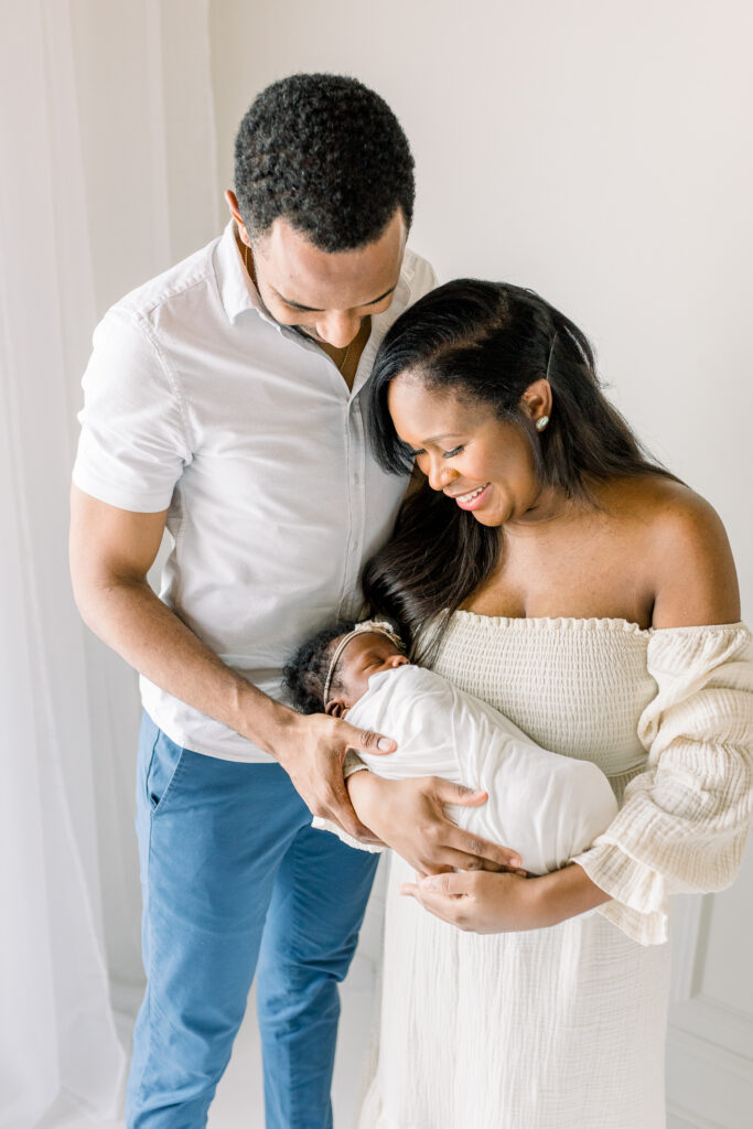 Newborn baby girl in white studio being held by mom and dad and mom and dad smiling at baby. Image by Mississippi Photographer.