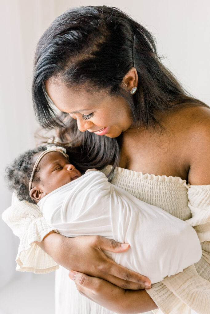 Newborn baby girl being held by mom in newborn white studio session. Mom is smiling down at baby in her arms. Image captured by Mississippi Newborn Photographer.