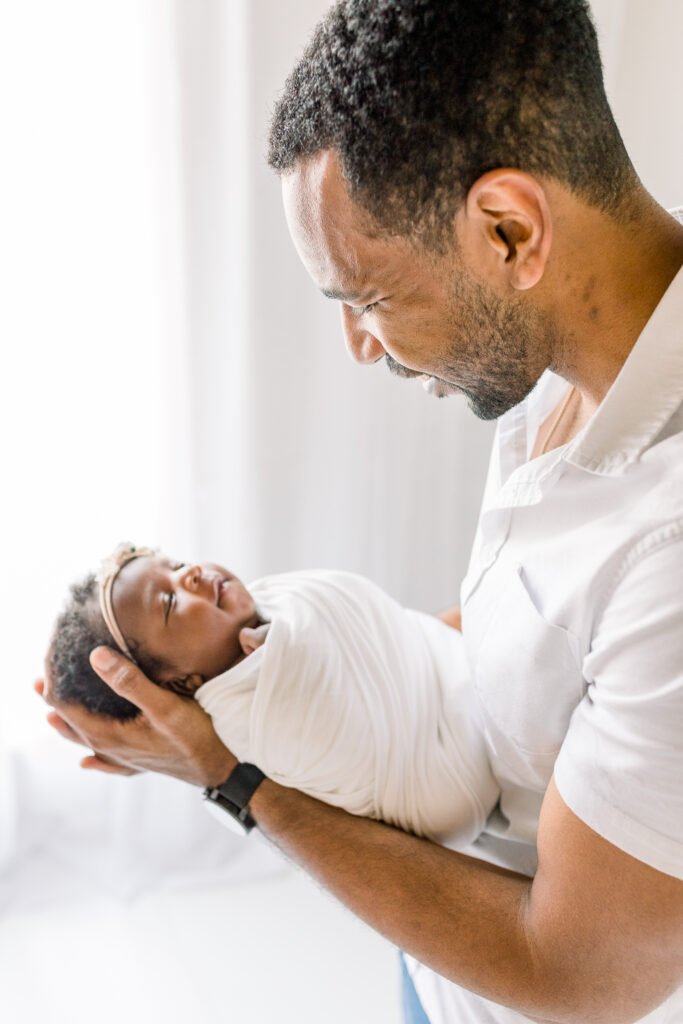 Newborn baby girl in white studio session with dad holding baby and smiling down at her. Image captured by Mississippi Newborn Photographer.