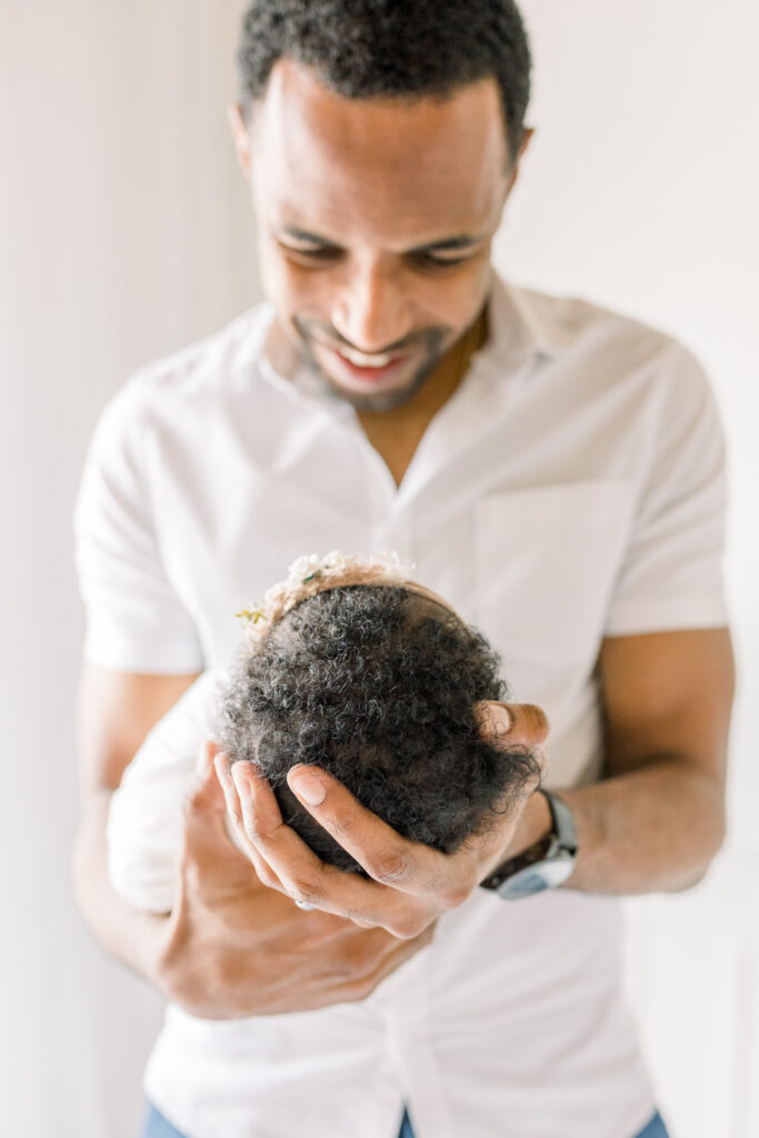 Dad holding baby smiling down at her in white studio. Close up on baby's hair. Image captured by Mississippi Newborn Photographer.