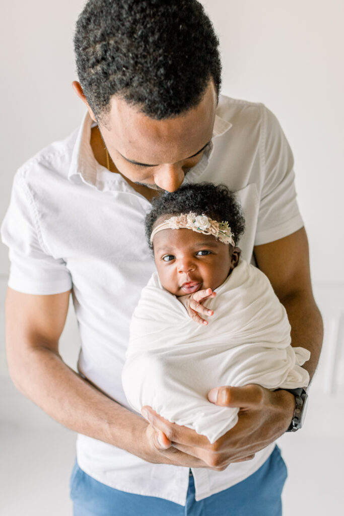 Dad holding newborn baby girl swaddled in white fabric smiling in white natural light studio by Mississippi Newborn Photographer. 