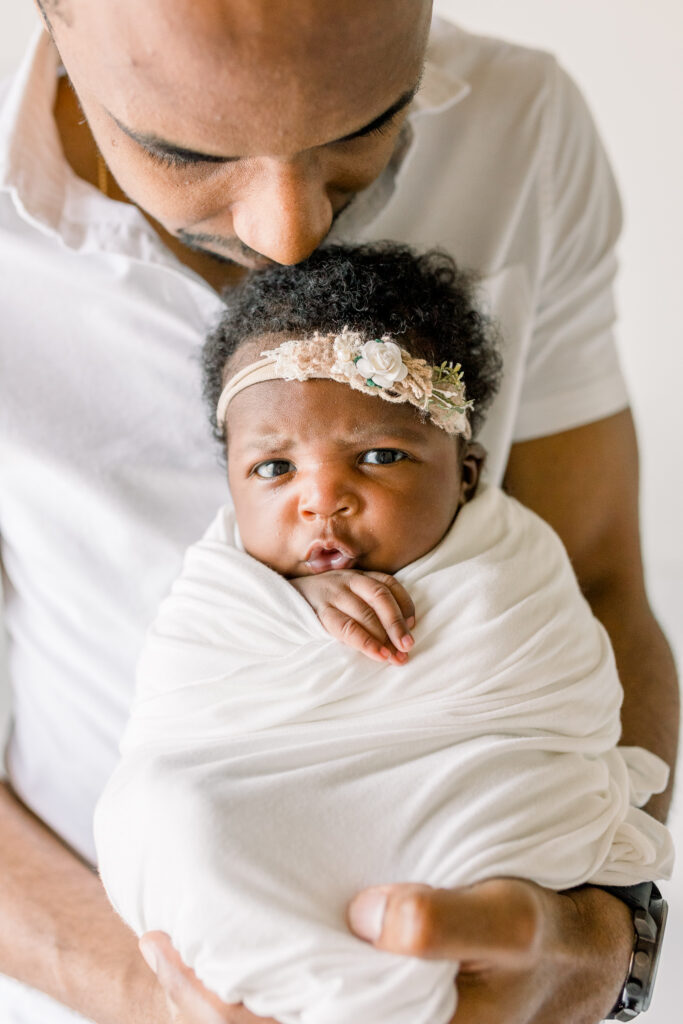 Newborn baby girl swaddled in white fabric making a face at camera while being held by dad. Image by Mississippi Newborn Photographer. 