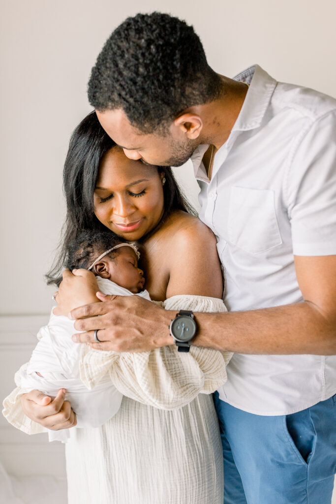 Newborn baby girl being held by mom and dad in white natural light studio. Dad kisses moms temple white mom smiles down at baby. Image by Mississippi Newborn Photographer. 