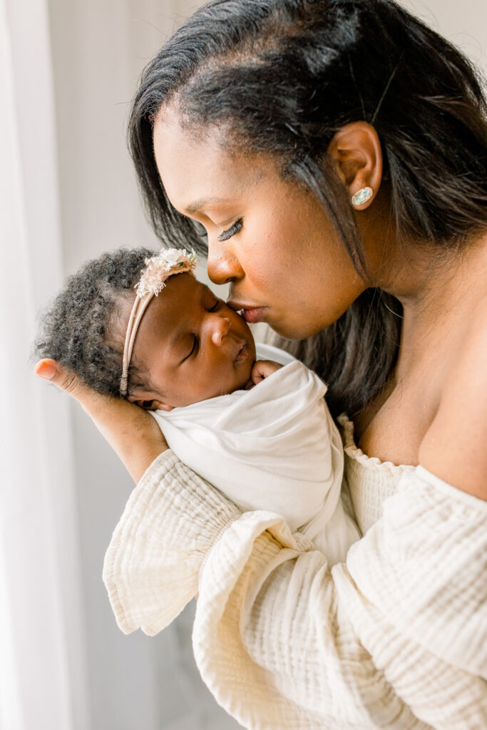 Newborn baby girl natural light studio session with mom holding baby and kissing her on the cheek. Image by Mississippi Newborn Photographer. 