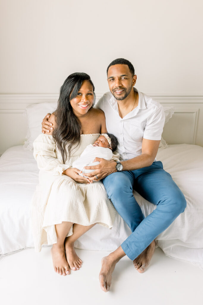 Mississippi Newborn Photographer captures image of mom and dad smiling at camera in natural light white studio while holding baby girl swaddled in white. 