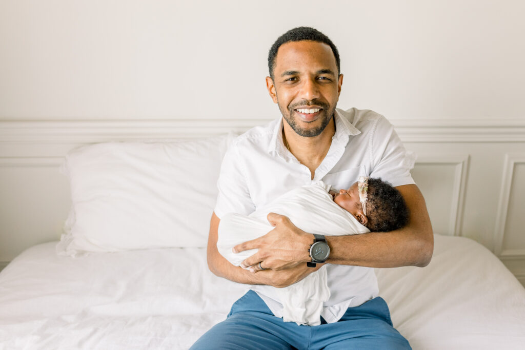 Dad sitting on white bed holding swaddled baby girl smiling at camera for studio newborn session by Mississippi Newborn Photographer. 