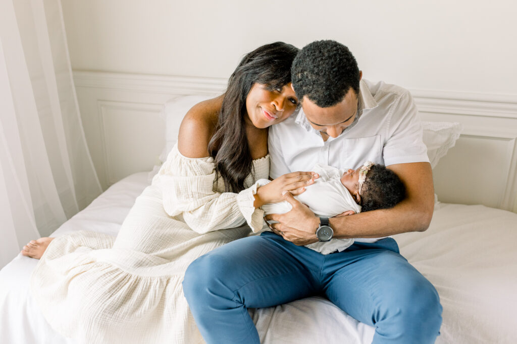 Mom and dad sitting on white bed in white natural light studio with mom smiling at camera and dad looking down at baby in natural light studio by Mississippi Newborn photographer. 