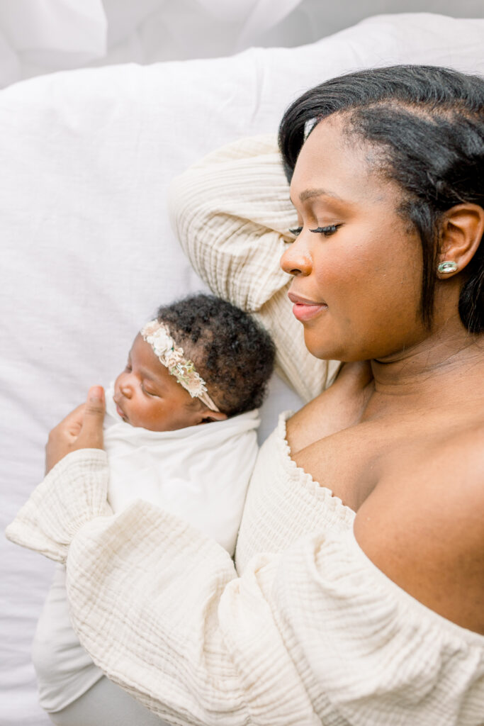 Mom lying on white bed holding newborn baby girl swaddled in white in natural light studio by Mississippi Newborn Photographer