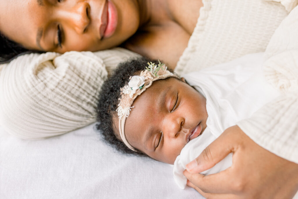 Mom lying on white bed holding newborn baby girl swaddled in white in natural light studio by Mississippi Newborn Photographer