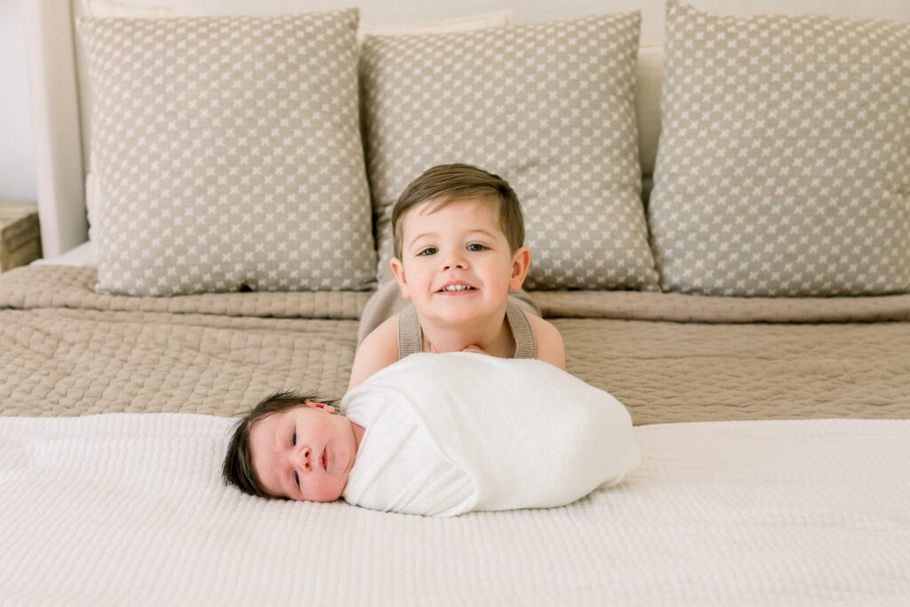 Toddler boy in neutral onsie smiling with swaddled newborn baby on master bed. Image taken by Madison, MS Newborn photographer.