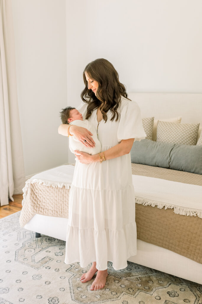 Mom standing in neutral toned master bed room holding swaddled newborn baby boy in white. Image taken by Madison, MS newborn photographer. 