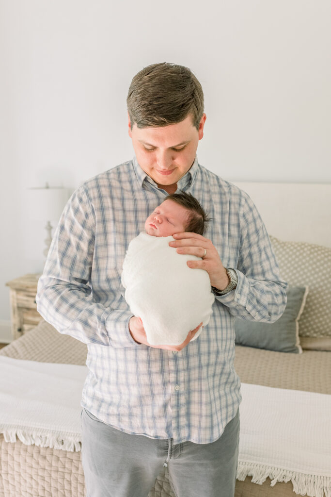 Dad wearing neutral toned plaid shirt holding swaddled baby and smiling down at baby. Image taken by CJ and Olive Photography, a Madison, MS photographer.