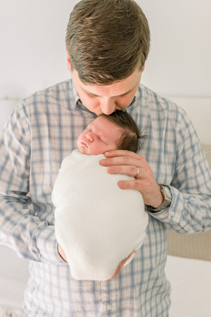 Dad wearing neutral toned plaid shirt holding swaddled baby and kissing the top of baby's head.  Image taken by CJ and Olive Photography, a Madison, MS photographer.