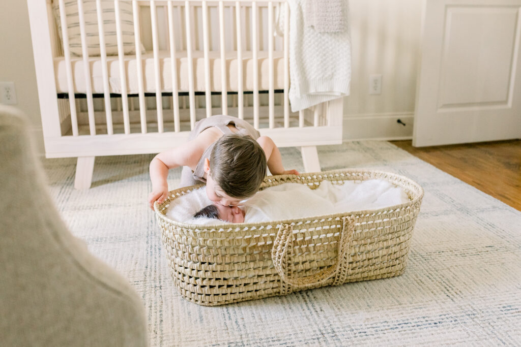 Swaddled newborn baby boy in moses basket in neutral toned nursery with toddler boy kissing baby. Image captured by Madison, MS newborn photographer.