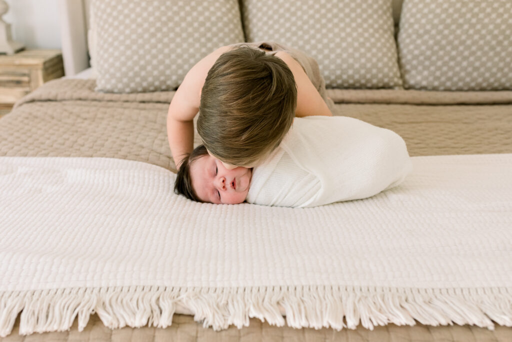 Little toddler boy dressed in neutral onesie kissing swaddled baby brother on neutral bedding. Image taken by Madison, MS newborn photographer. 