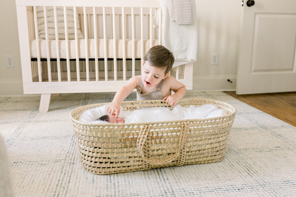 Swaddled newborn baby boy in moses basket in neutral toned nursery with toddler boy pointing to baby. Image captured by Madison, MS newborn photographer.