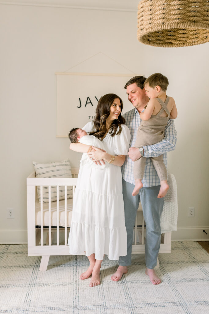 Family standing together in front of crib in neutral toned nursery while holding swaddled newborn baby boy in white. Image taken by Madison, MS newborn photographer.