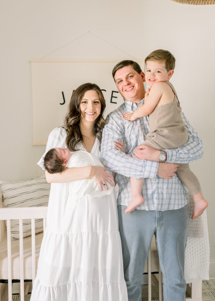 Family standing together in front of crib in neutral toned nursery while holding swaddled newborn baby boy in white. Image taken by Madison, MS newborn photographer.