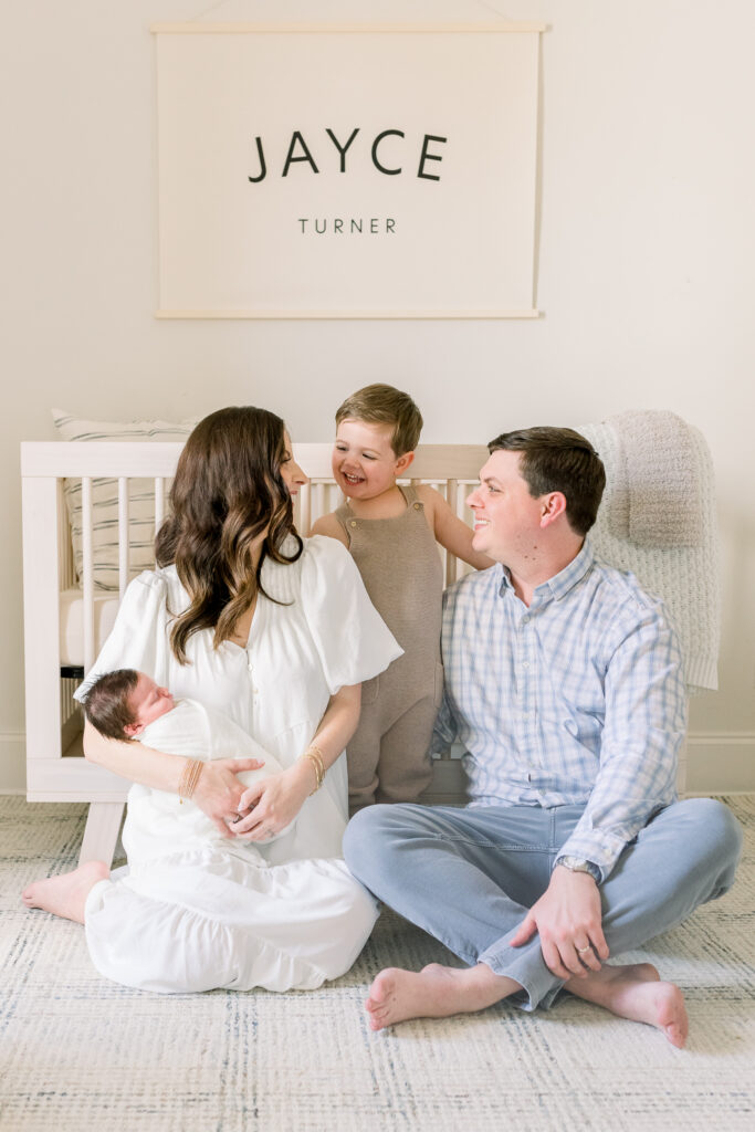 Family sitting together in front of crib in neutral toned nursery while holding swaddled newborn baby boy in white. Image taken by Madison, MS newborn photographer.