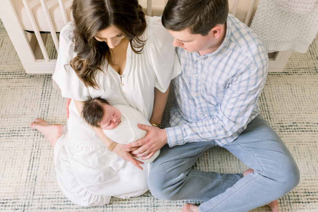 Mom and dad sitting together in front of crib holding swaddled newborn baby boy in white. Image shot from above by CJ and Olive Photography, a Madison, MS Newborn photographer. 