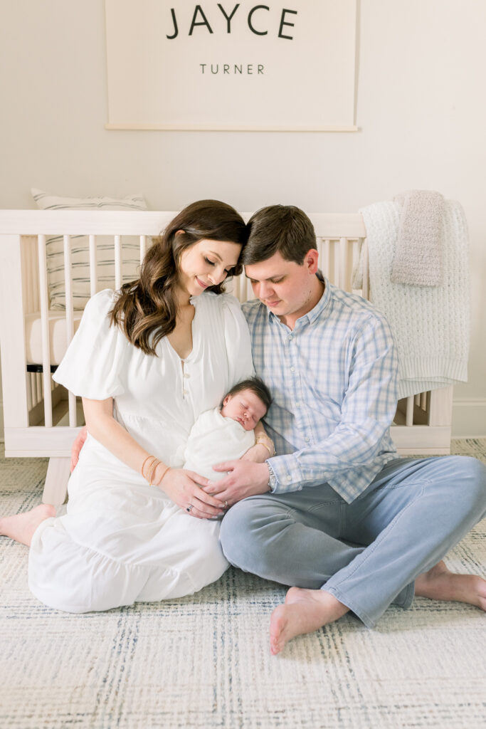 Mom and dad sitting together in front of crib holding swaddled newborn baby boy in white. Image taken by CJ and Olive Photography, a Madison, MS Newborn photographer. 