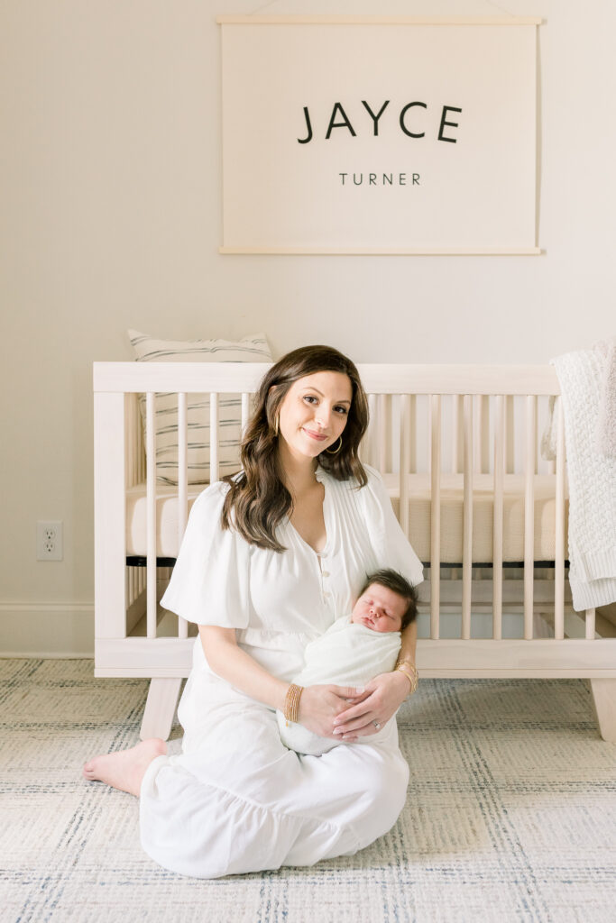 Mom sitting in front of crib holding swaddled newborn baby boy in white. Image taken by CJ and Olive Photography, a Madison, MS Newborn photographer. 