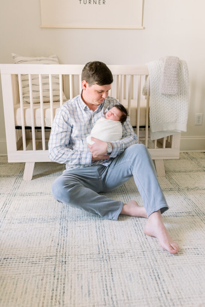 Dad sitting in front of crib holding swaddled newborn baby boy in white. Image taken by CJ and Olive Photography, a Madison, MS Newborn photographer. 