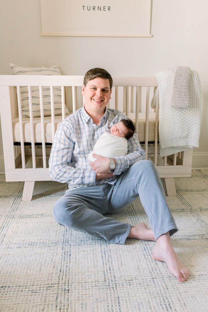 Dad sitting in front of crib holding swaddled newborn baby boy in white. Image taken by CJ and Olive Photography, a Madison, MS Newborn photographer. 