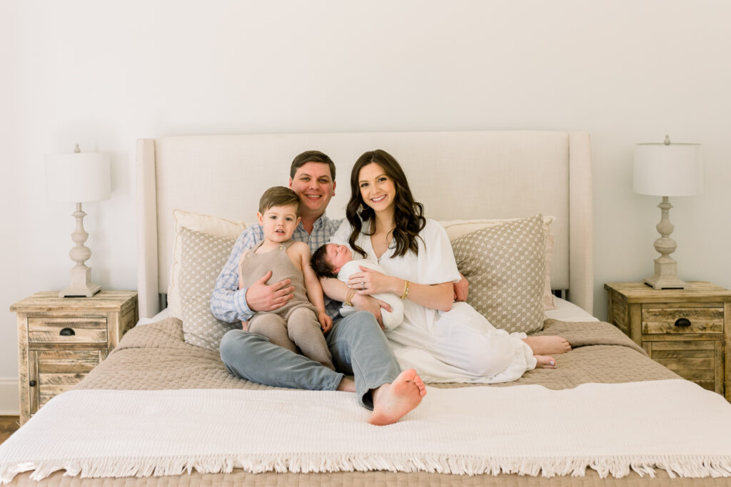 Family smiling together at camera while holding swaddled baby boy in white in a neutral toned master bedroom. Image captured by Madison, MS newborn photographer.