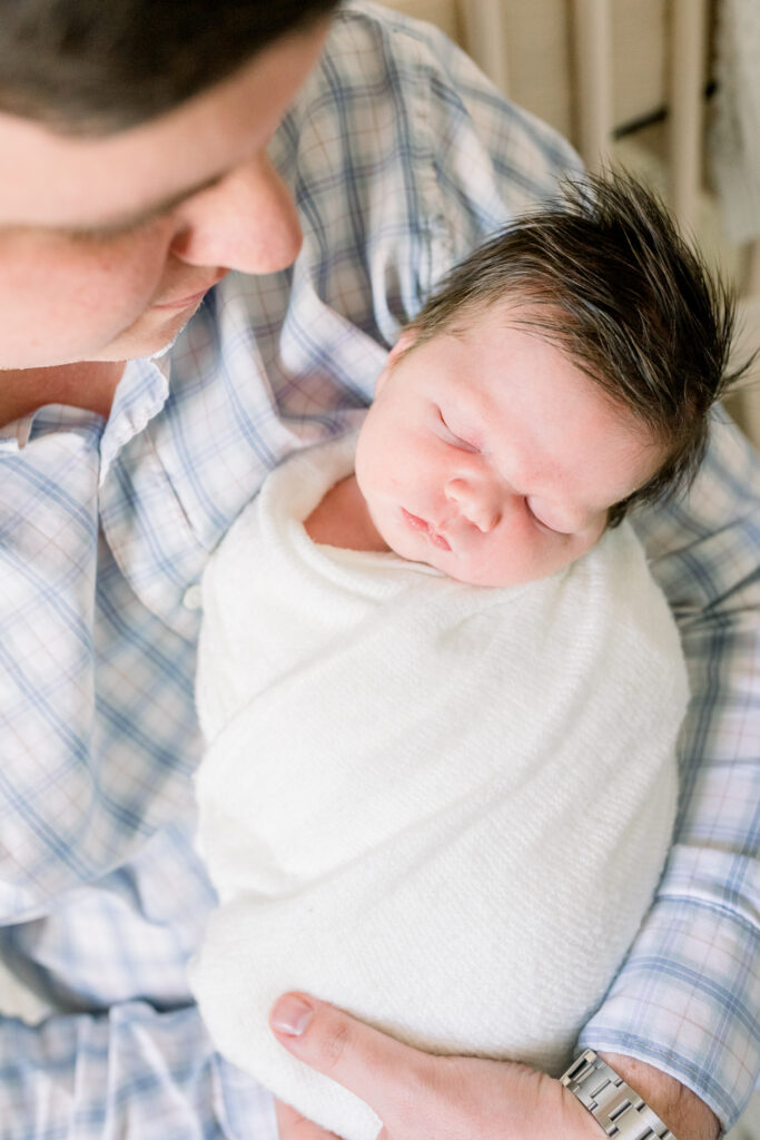 Dad sitting in front of crib holding swaddled newborn baby boy in white. Image is a close up on baby in dad's arms by CJ and Olive Photography, a Madison, MS Newborn photographer. 