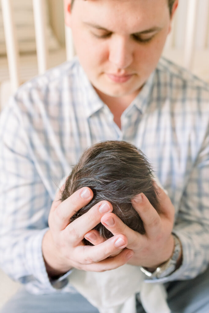 Dad sitting in front of crib holding swaddled newborn baby boy in white. Image taken by CJ and Olive Photography, a Madison, MS Newborn photographer. 