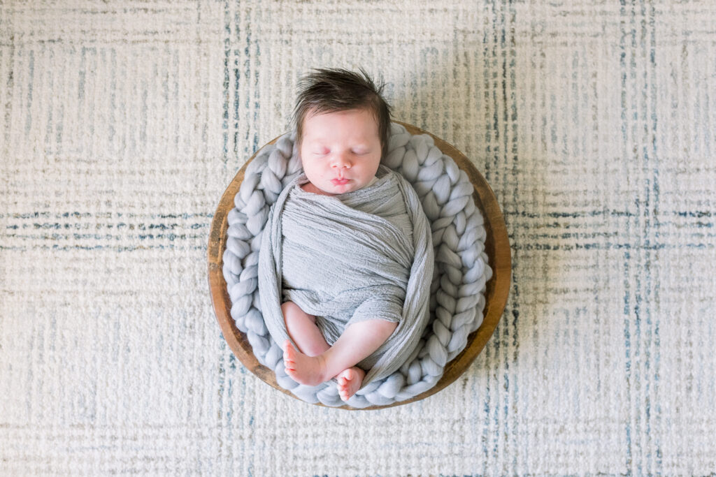 Newborn baby boy swaddled in grey in wooden bowl with grey stuffing. Image captured by Madison, MS newborn photographer, CJ and Olive Photography.