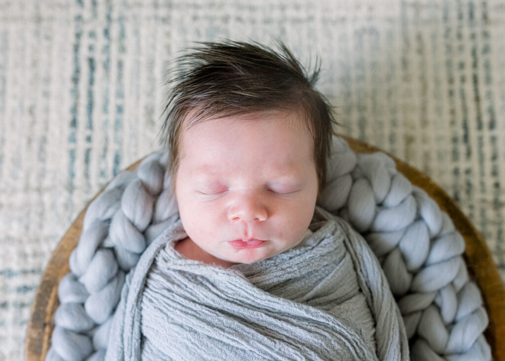 Newborn baby boy swaddled in grey in wooden bowl with grey stuffing. Image captured by Madison, MS newborn photographer, CJ and Olive Photography.