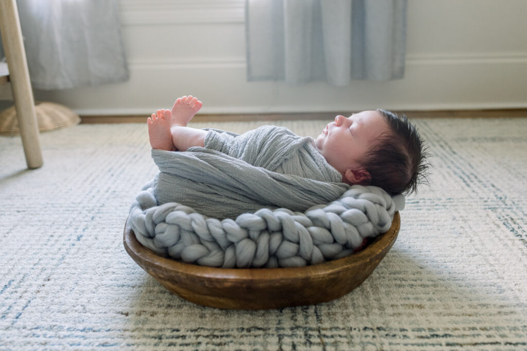 Profile shot of newborn baby boy swaddled in grey in wooden bowl with grey stuffing. Image captured by Madison, MS newborn photographer, CJ and Olive Photography.