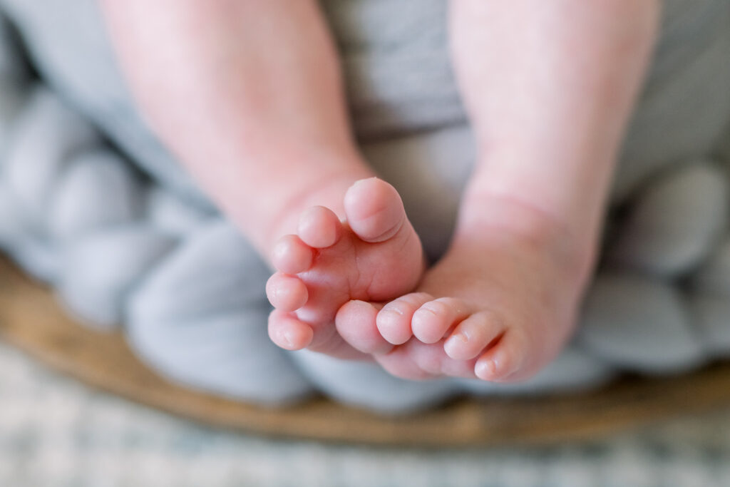 Close up on newborn baby boy's toes swaddled in grey in wooden bowl with grey stuffing. Image captured by Madison, MS newborn photographer, CJ and Olive Photography.