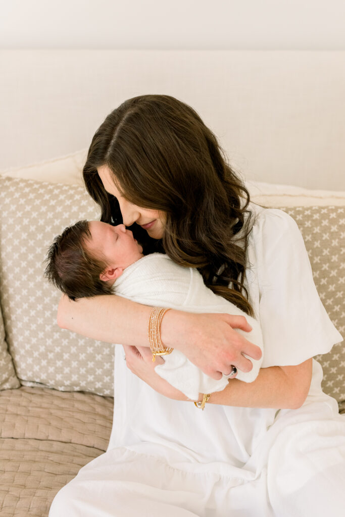 Mom sitting on neutral colored bedding  nose to nose with swaddled baby boy. Image captured by CJ and Olive photography, a Madison, MS Newborn photographer. 