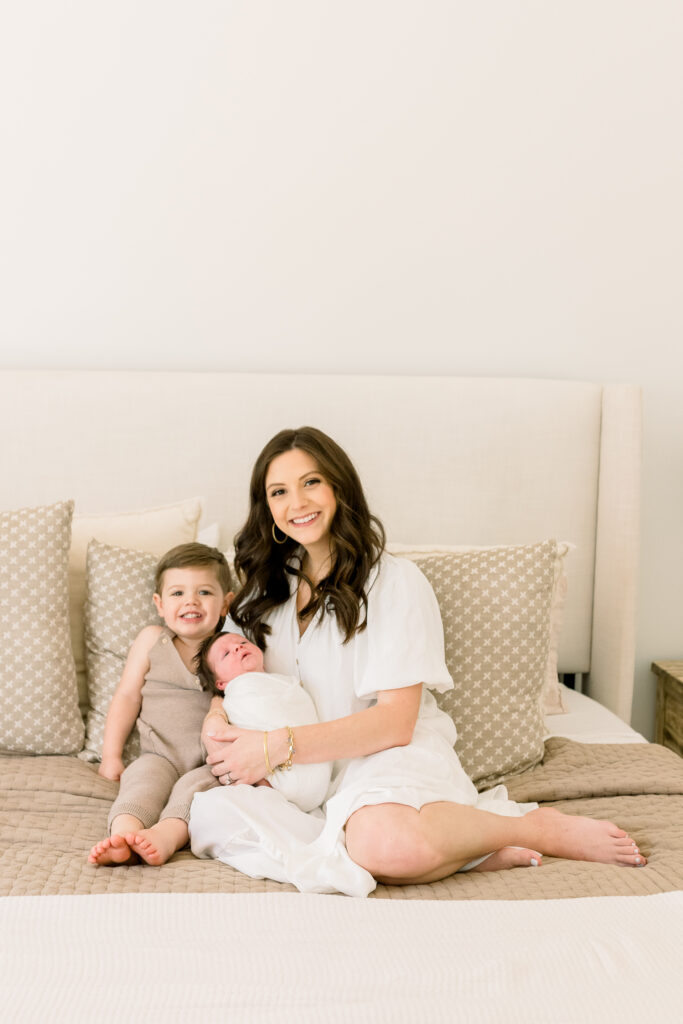 Mom posing with toddler boy and swaddled newborn baby boy smiling at camera in neutral toned master bedroom. Image captured by Madison newborn photographer.