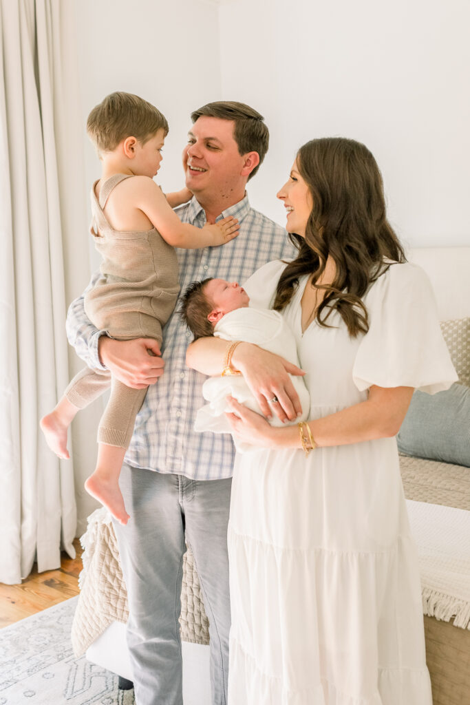 Family standing in neutral toned bedroom smiling at each other while holding swaddled newborn baby boy. Image taken by Madison, MS newborn photographer. 