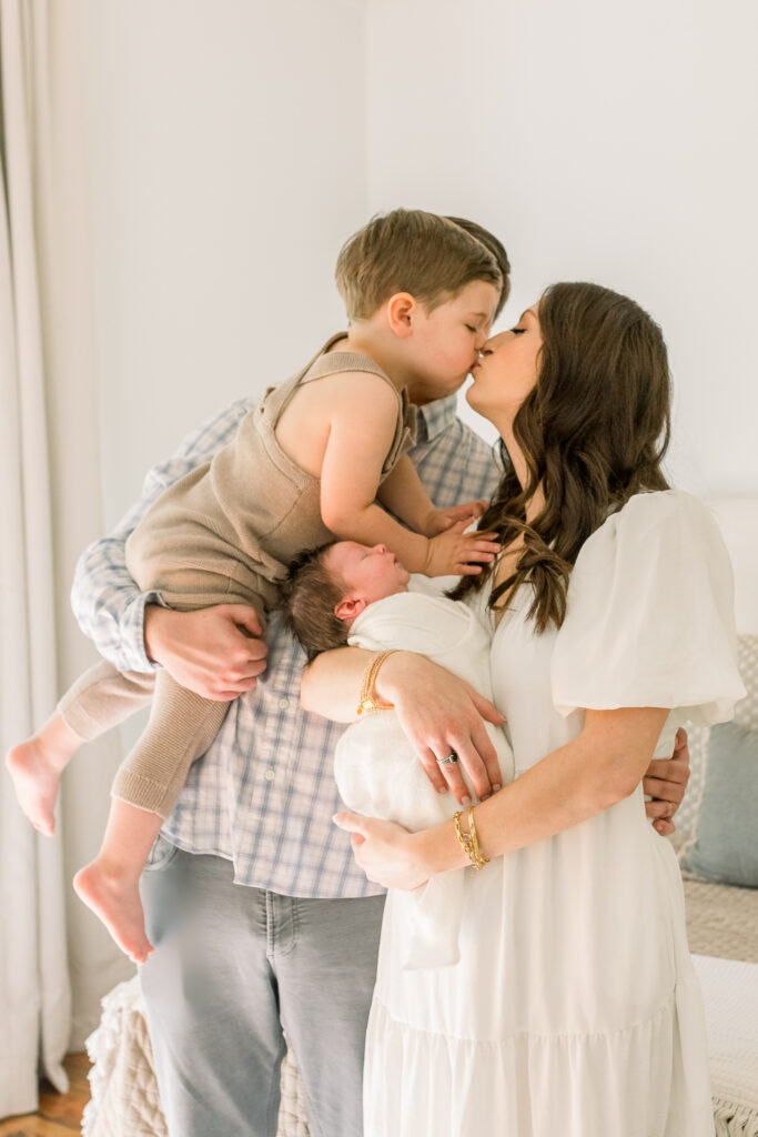 Family standing in neutral toned bedroom with mom kissing toddler boy and holding swaddled newborn baby boy. Image taken by Madison, MS newborn photographer. 