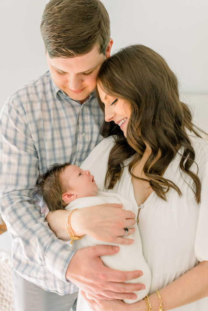 Close up image of mom and dad smiling at swaddled baby boy with beautiful soft natural light on them. Image captured by Madison, MS photographer CJ and Olive photography. 