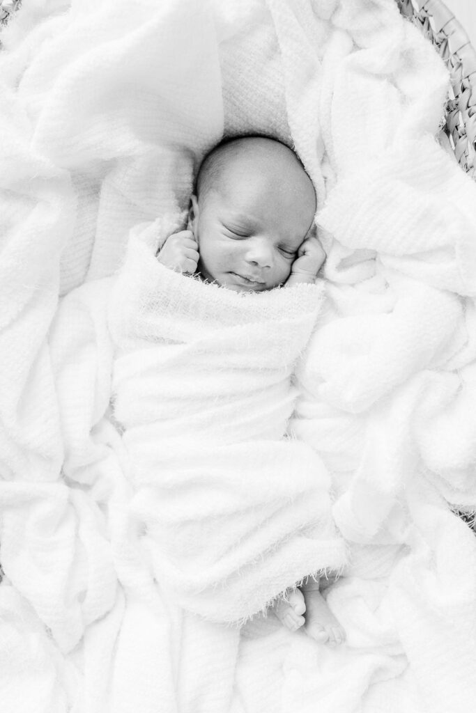 A black and white image of newborn baby boy swaddled in white in Moses basket in white studio by CJ and Olive Photography, a Brandon newborn photographer. 