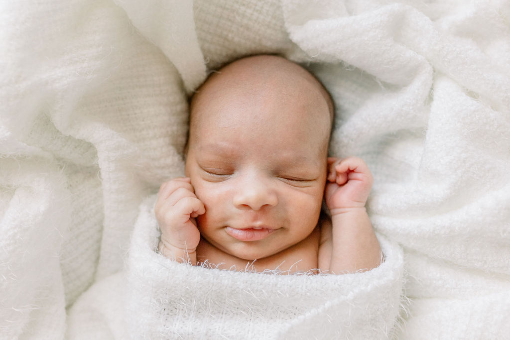 Newborn baby boy smiles while swaddled in white in Moses basket during newborn studio session. Image taken by Brandon newborn photographer. 