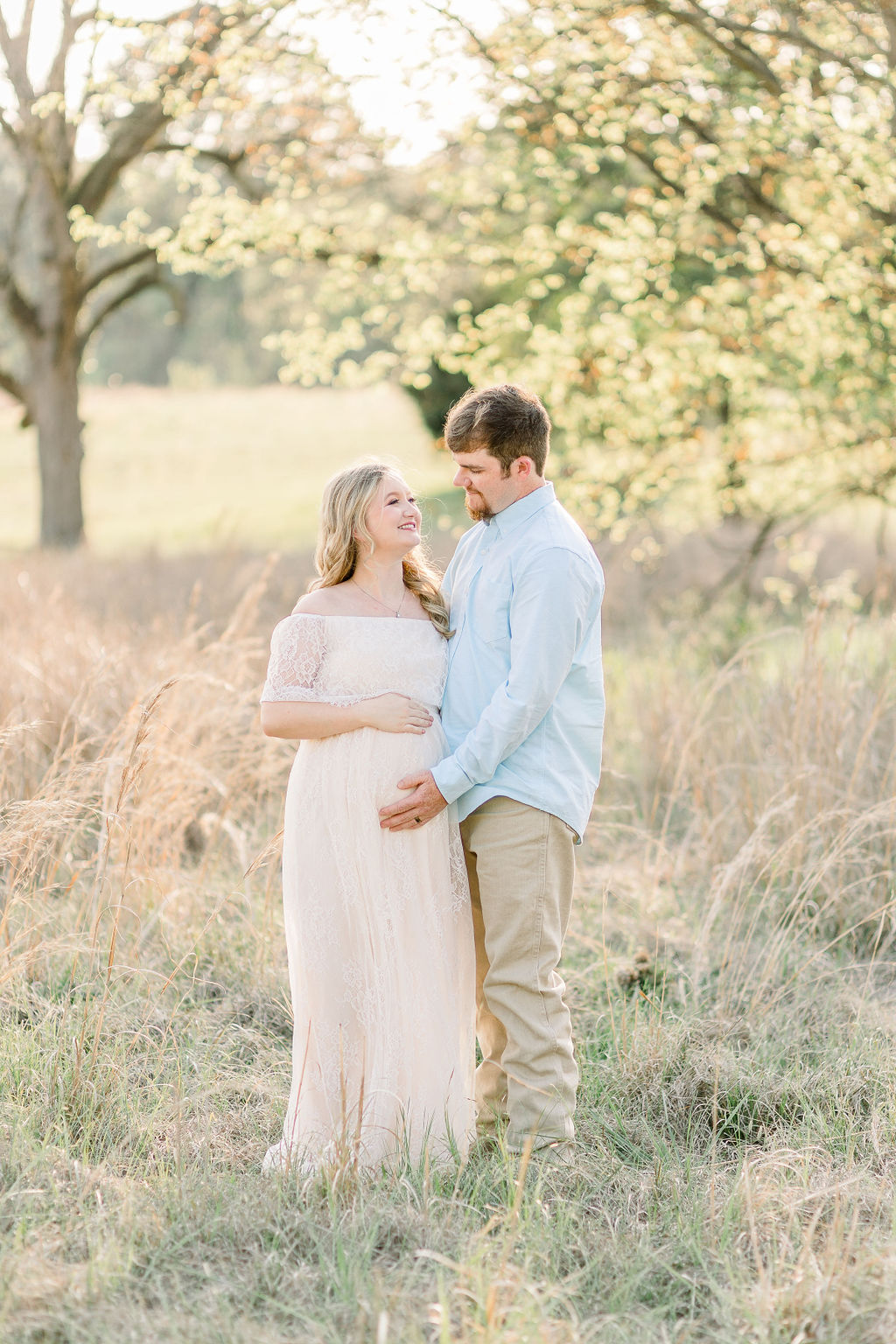 Mom and dad to be smiling at each other in green field with beautiful back lighting. Image captured by Madison Maternity Photographer.