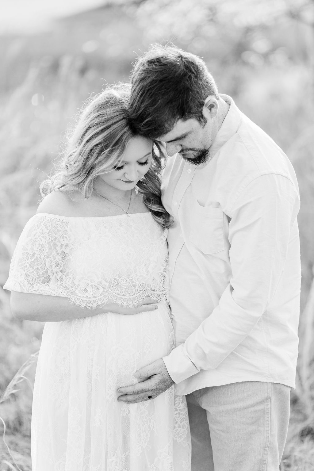 Mom and dad pose for maternity session in field while looking down at the belly. Black and white Image captured by Madison maternity photographer. 