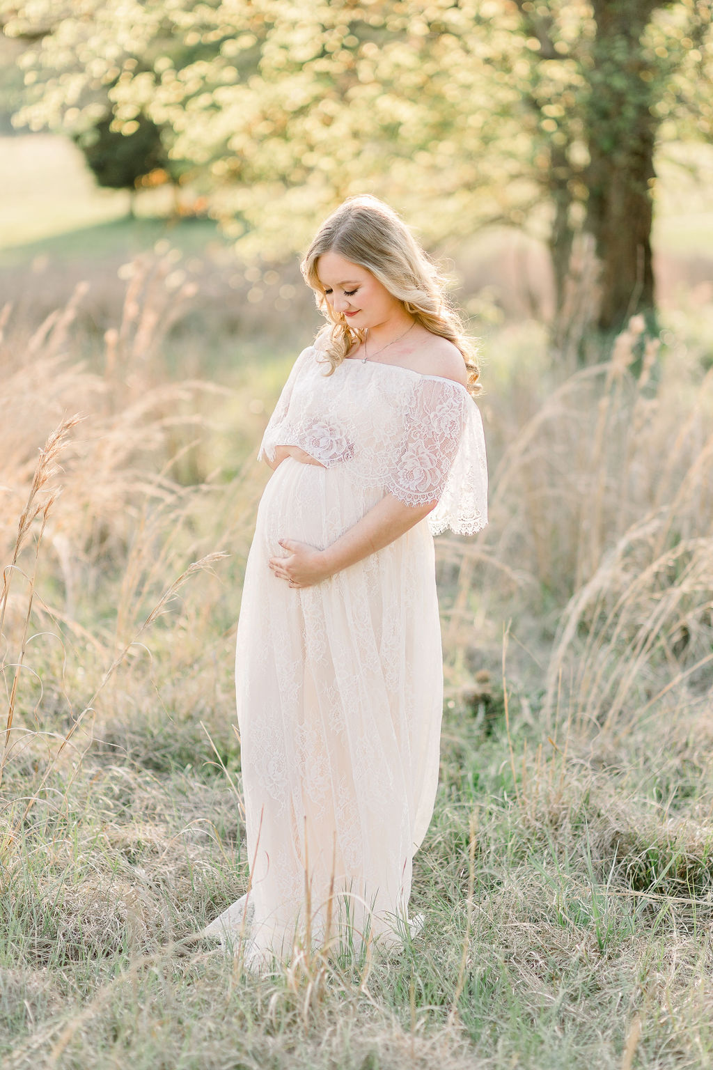 Mom to be smiles down at belly while wearing white lace maxi dress in a green field at sunset. Image captured by CJ and Olive Photography, a Madison, Mississippi maternity photographer. 