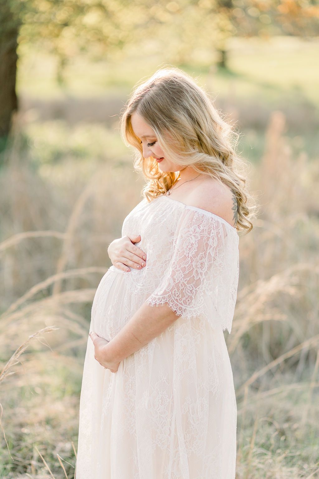 Mom wears white lace maxi dress in field with beautiful rim light lighting behind her while she smiles down at belly. Image captured by CJ and Olive Photography, a Madison maternity photographer.